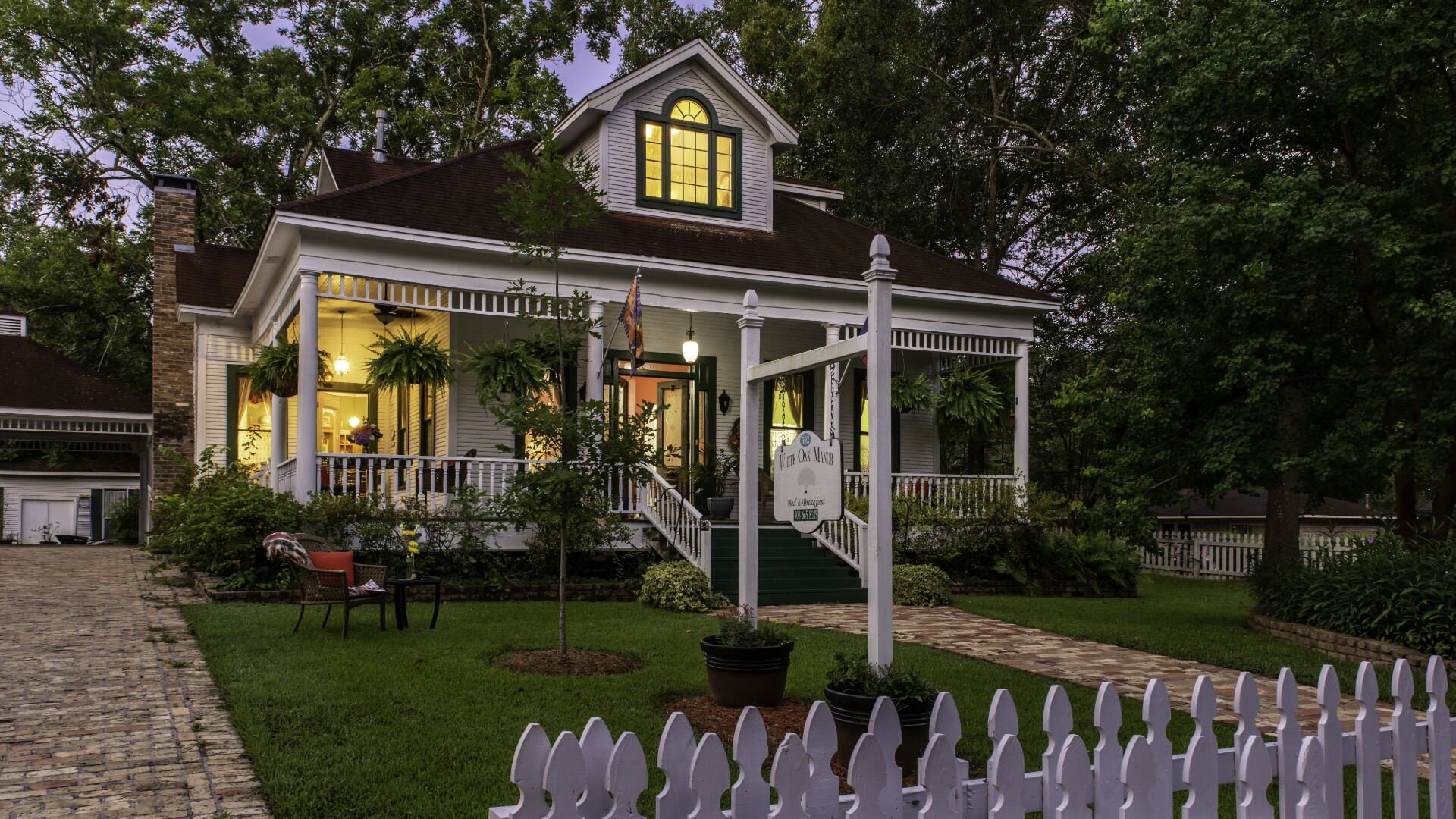 Exterior view of a property painted white with white trim, wrapped front porch, white picket fence, green grass, and surrounded by large green trees
