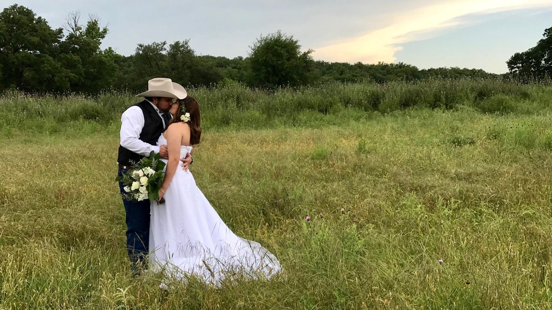 Groom holding and kissing bride while standing in field of grass with trees in the background