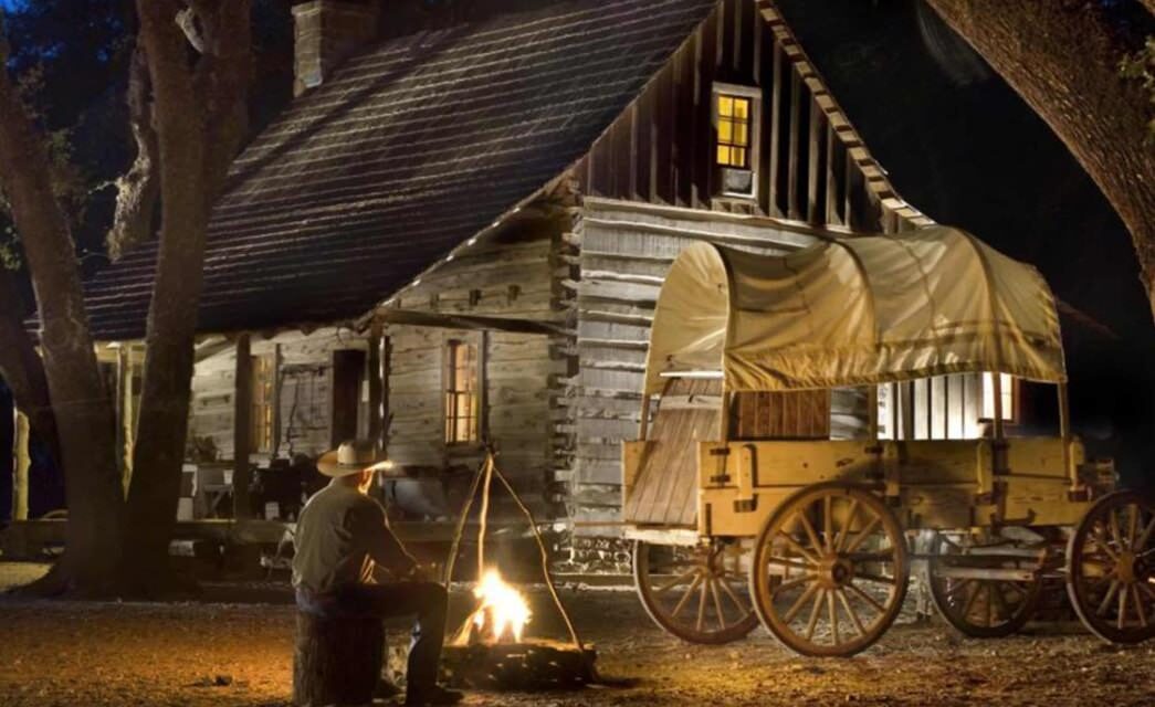 Cowboy sitting on stump next to firepit and old antique pioneer wagon with log cabin in background at night